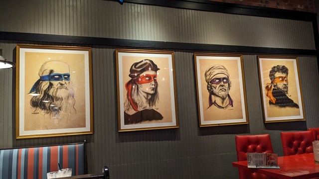Four framed sketches hang on a wall behind tables at a chain table service restaurant. Each sketch is of the head of a Renaissance artist, in black pencil with the only color being a bandana with eye holes cut out. From left to right: Leonardo Da Vinci is wearing blue, Raphael wearing red, Donatello wearing purple, and Michelangelo wearing orange.