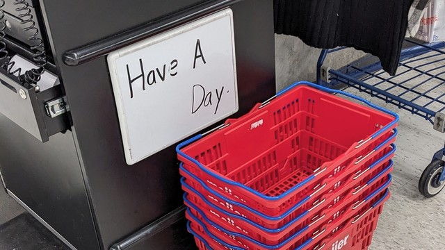 A stack of red shopping baskets sit in front of a gray stand in a store. A white board is attached to the gray stand. Someone has written these words: Have a day. There is a blank space between the word a and the word day.