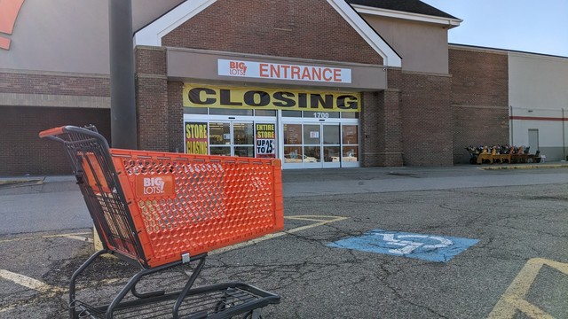 An empty orange shopping cart with the words Big Lots sits in a  parking lot in front of a shop. A sign above the wide sliding entrance doors reads "Big Lots Entrance." Between that sign and the sliding doors is a vinyl banner that reads "Closing" in huge letters. Two big signs are posted on the doors. One reads "store closing" and the other reads "Entire store up to 25% off." The pavement of the parking lot has numerous cracks.