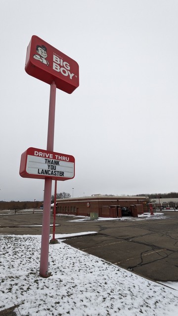 A sign for the restaurant Frisch's Big Boy looms high in a cloudy sky on a pole. The sign features a smiling cartoon boy with a pompadour haircut. Halfway up the pole is a second sign that reads "Drive thru" and "Thank you Lancaster". Behind the sign is an empty parking lot and a deserted one-story brick building. The parking lot has faded yellow lines for parking spots and lots of tar patching cracks in the asphalt. The parking lot is surrounded by grass with a light dusting of snow.